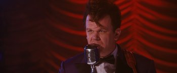 Movie still from “Walk Hard: The Dewey Cox Story” (2007), directed by Jake Kasdan – A man in a suit and bow tie is singing into a microphone; Close Up shot, Over the shoulder angle