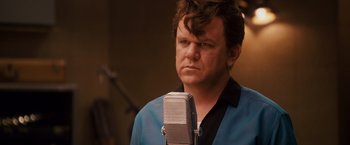 Movie still from “Walk Hard: The Dewey Cox Story” (2007), directed by Jake Kasdan – A man in a blue shirt is standing in front of a microphone; Close Up shot, Over the shoulder angle