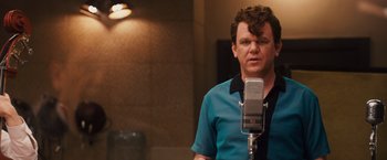 Movie still from “Walk Hard: The Dewey Cox Story” (2007), directed by Jake Kasdan – A man standing in front of a microphone in a room; Close Up shot, Over the shoulder angle