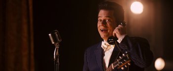 Movie still from “Walk Hard: The Dewey Cox Story” (2007), directed by Jake Kasdan – A man in a suit talking on a phone; Medium shot, Low angle