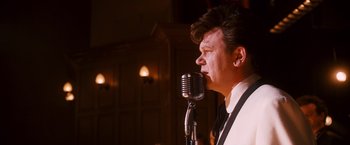 Movie still from “Walk Hard: The Dewey Cox Story” (2007), directed by Jake Kasdan – A man in a white shirt and black tie standing next to a microphone; Close Up shot, Over the shoulder angle