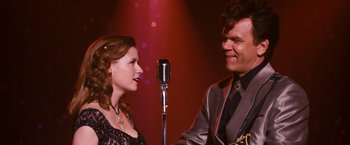 Movie still from “Walk Hard: The Dewey Cox Story” (2007), directed by Jake Kasdan – A man and a woman sing into microphones in front of a microphone stand; Medium shot, Over the shoulder angle
