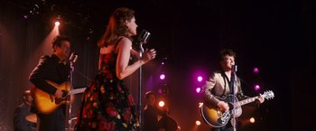 Movie still from “Walk Hard: The Dewey Cox Story” (2007), directed by Jake Kasdan – A woman in a floral dress singing into a microphone while a man plays guitar; Medium shot, Over the shoulder angle
