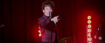 Movie still from “Walk Hard: The Dewey Cox Story” (2007), directed by Jake Kasdan – A man holding a microphone and pointing to the side; Medium shot, Over the shoulder angle