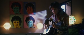 Movie still from “Walk Hard: The Dewey Cox Story” (2007), directed by Jake Kasdan – A man playing a guitar in front of a wall of pictures; Medium shot, Low angle