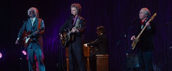 Movie still from “Walk Hard: The Dewey Cox Story” (2007), directed by Jake Kasdan – A man in a suit and tie playing a guitar and a piano; Medium shot, Over the shoulder angle