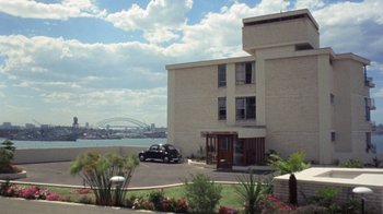 Movie still from “Walkabout” (1971), directed by Nicolas Roeg – A car parked in front of a building near a body of water; Extreme Wide shot, High angle