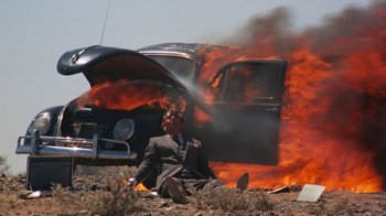 Movie still from “Walkabout” (1971), directed by Nicolas Roeg – A man sitting in the grass near a car on fire; Wide shot, Low angle