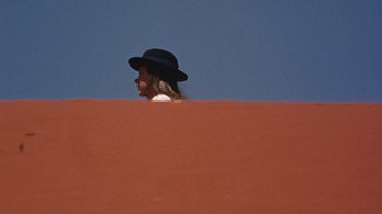Movie still from “Walkabout” (1971), directed by Nicolas Roeg – A woman wearing a black hat is leaning on a red wall; Wide shot, Overhead angle