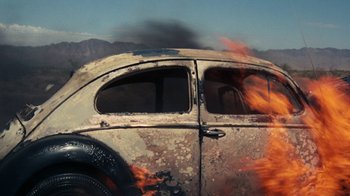 Movie still from “Walkabout” (1971), directed by Nicolas Roeg – An old car is on fire in the middle of the road; Wide shot, Overhead angle