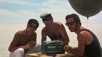 Movie still from “Walkabout” (1971), directed by Nicolas Roeg – A group of men sitting next to each other on the beach; Medium shot, Low angle