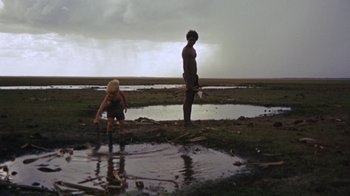 Movie still from “Walkabout” (1971), directed by Nicolas Roeg – Two people standing in a puddle of water near a body of water; Wide shot, High angle