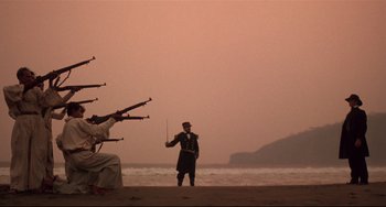 Movie still from “Walker” (1987), directed by Alex Cox – A man standing on a beach next to a bunch of guns; Wide shot, Low angle