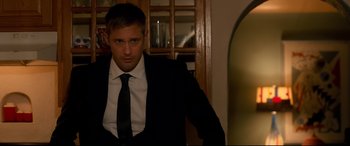 Movie still from “War on Everyone” (2016), directed by John Michael McDonagh – A man in a suit and tie standing in front of a window; Medium shot, Over the shoulder angle