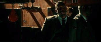 Movie still from “War on Everyone” (2016), directed by John Michael McDonagh – A man wearing a suit and tie standing in front of a wooden wall; Medium shot, Over the shoulder angle