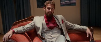 Movie still from “War on Everyone” (2016), directed by John Michael McDonagh – A man sitting on a chair wearing a suit and tie; Medium shot, Low angle