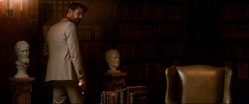Movie still from “War on Everyone” (2016), directed by John Michael McDonagh – A man standing next to a bust in a library; Medium shot, Over the shoulder angle