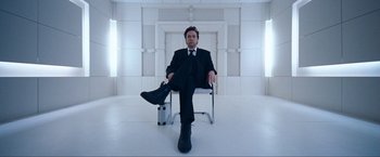 Movie still from “War, Inc.” (2008), directed by Joshua Seftel – A man in a suit and tie sitting on a chair; Wide shot, Low angle