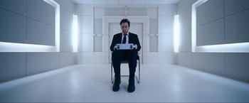 Movie still from “War, Inc.” (2008), directed by Joshua Seftel – A man in a suit and tie sitting in a chair; Wide shot, Overhead angle