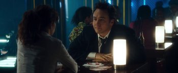 Movie still from “War, Inc.” (2008), directed by Joshua Seftel – A man sitting at a table in front of a light; Medium shot, Over the shoulder angle