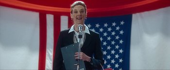 Movie still from “War, Inc.” (2008), directed by Joshua Seftel – A woman standing at a podium holding a microphone; Medium shot, Low angle