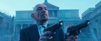 Movie still from “War, Inc.” (2008), directed by Joshua Seftel – An older man in a suit and tie holding a gun; Close Up shot, Low angle