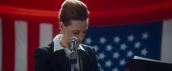 Movie still from “War, Inc.” (2008), directed by Joshua Seftel – A woman in a black jacket is singing into a microphone; Medium shot, Low angle