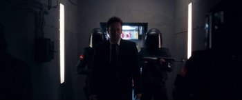 Movie still from “War, Inc.” (2008), directed by Joshua Seftel – A man in a suit standing in front of a group of men; Medium shot, Low angle