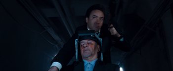 Movie still from “War, Inc.” (2008), directed by Joshua Seftel – A man in a suit and a hat is sitting in a chair with another man in a suit; Close Up shot, Low angle