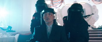 Movie still from “War, Inc.” (2008), directed by Joshua Seftel – An older man wearing a suit and a hat; Medium shot, Over the shoulder angle