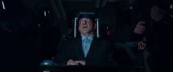 Movie still from “War, Inc.” (2008), directed by Joshua Seftel – An older man in a suit and tie; Medium shot, Over the shoulder angle