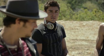 Movie still from “We Are Your Friends” (2015), directed by Max Joseph – A young man wearing headphones and smoking a cigarette while standing next to another man; Medium shot, Over the shoulder angle