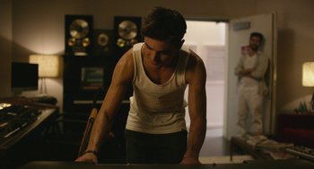 Movie still from “We Are Your Friends” (2015), directed by Max Joseph – A man in a white tank top and black pants is standing in front of a table; Medium shot, Over the shoulder angle