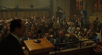 Movie still from “We Own the Night” (2007), directed by James Gray – A group of people sitting in front of a microphone in a room; Wide shot, High angle