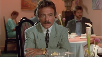 Movie still from “Weekend at Bernie's” (1989), directed by Ted Kotcheff – A man sitting at a dinner table with a watch; Medium shot, Over the shoulder angle