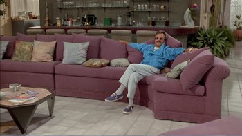 Movie still from “Weekend at Bernie's” (1989), directed by Ted Kotcheff – A man sitting on top of a pink couch in a living room; Wide shot, High angle