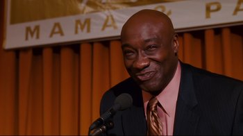 Movie still from “Welcome Home, Roscoe Jenkins” (2008), directed by Malcolm D. Lee – A man wearing a suit and tie smiles at a microphone; Close Up shot, High angle