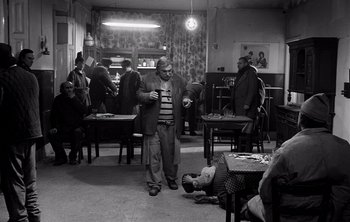 Movie still from “Werckmeister Harmonies” (2000), directed by Ágnes Hranitzky – An older man standing next to a group of people in a room; Wide shot, High angle