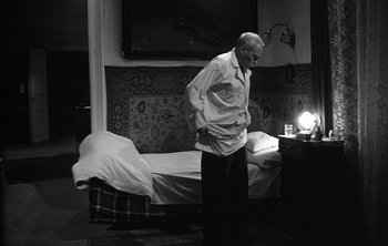 Movie still from “Werckmeister Harmonies” (2000), directed by Ágnes Hranitzky – An older man standing in front of a bed in a room; Wide shot, High angle