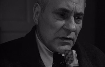 Movie still from “Werckmeister Harmonies” (2000), directed by Ágnes Hranitzky – An older man in a suit and tie holding a microphone; Close Up shot, Low angle