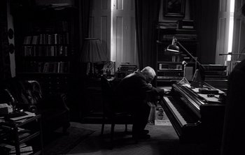 Movie still from “Werckmeister Harmonies” (2000), directed by Ágnes Hranitzky – An older man sitting in a chair in front of a piano; Wide shot, High angle