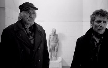 Movie still from “Werckmeister Harmonies” (2000), directed by Ágnes Hranitzky – A man standing next to another man in a bathroom; Medium shot, Over the shoulder angle