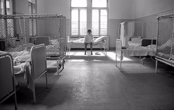 Movie still from “Werckmeister Harmonies” (2000), directed by Ágnes Hranitzky – A child sitting on a bed in a room with several beds in it; Wide shot, High angle