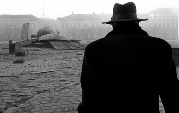 Movie still from “Werckmeister Harmonies” (2000), directed by Ágnes Hranitzky – A man wearing a hat and a black coat is standing on a cobblestone street; Wide shot, Low angle