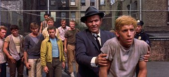 Movie still from “West Side Story” (1961), directed by Jerome Robbins – A man wearing a suit and a hat standing in front of a group of young men; Medium shot, Over the shoulder angle