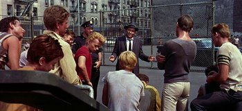 Movie still from “West Side Story” (1961), directed by Jerome Robbins – A group of people gathered around a man in a suit and tie; Medium shot, Over the shoulder angle