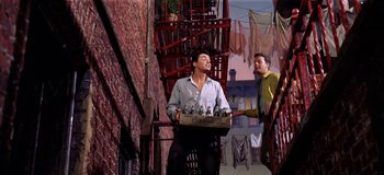 Movie still from “West Side Story” (1961), directed by Jerome Robbins – A man holding a crate of bottles while another man watches; Medium shot, Low angle