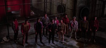 Movie still from “West Side Story” (1961), directed by Jerome Robbins – A group of young men standing next to each other in front of a chain link fence; Wide shot, High angle