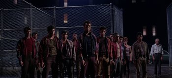 Movie still from “West Side Story” (1961), directed by Jerome Robbins – A group of men standing next to each other in front of a chain link fence; Wide shot, Low angle