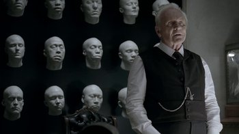 Movie still from “Westworld” (2016), created by Lisa Joy – A man in a suit and tie standing in front of a wall of heads; Close Up shot, Over the shoulder angle